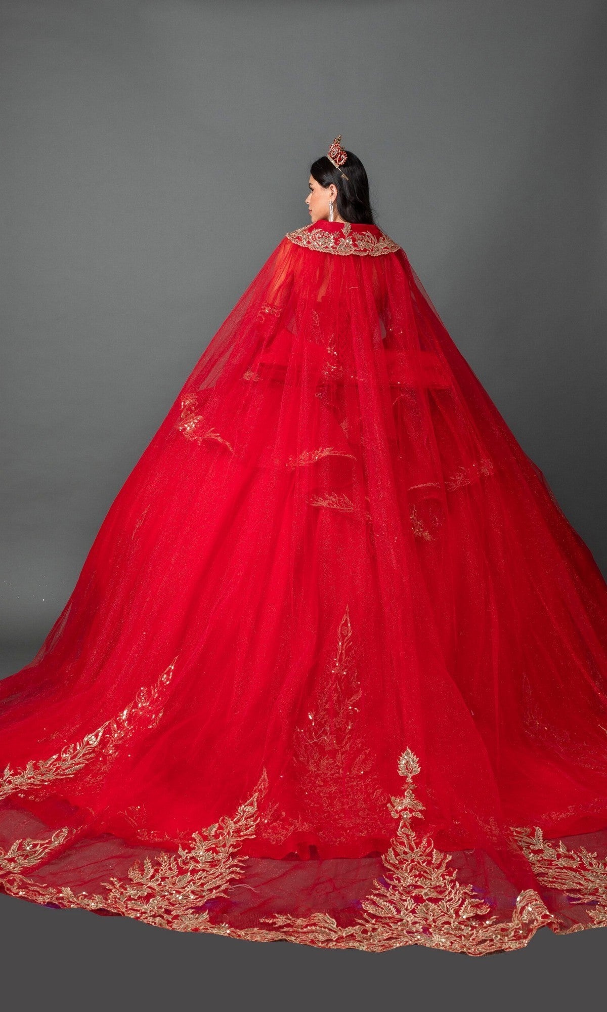 A woman stands facing away against a plain gray background, wearing the Quinceanera Dress 5161 By Dancing Queen—a red strapless ballgown featuring gold embroidered appliques and a long train—and a matching red crown.